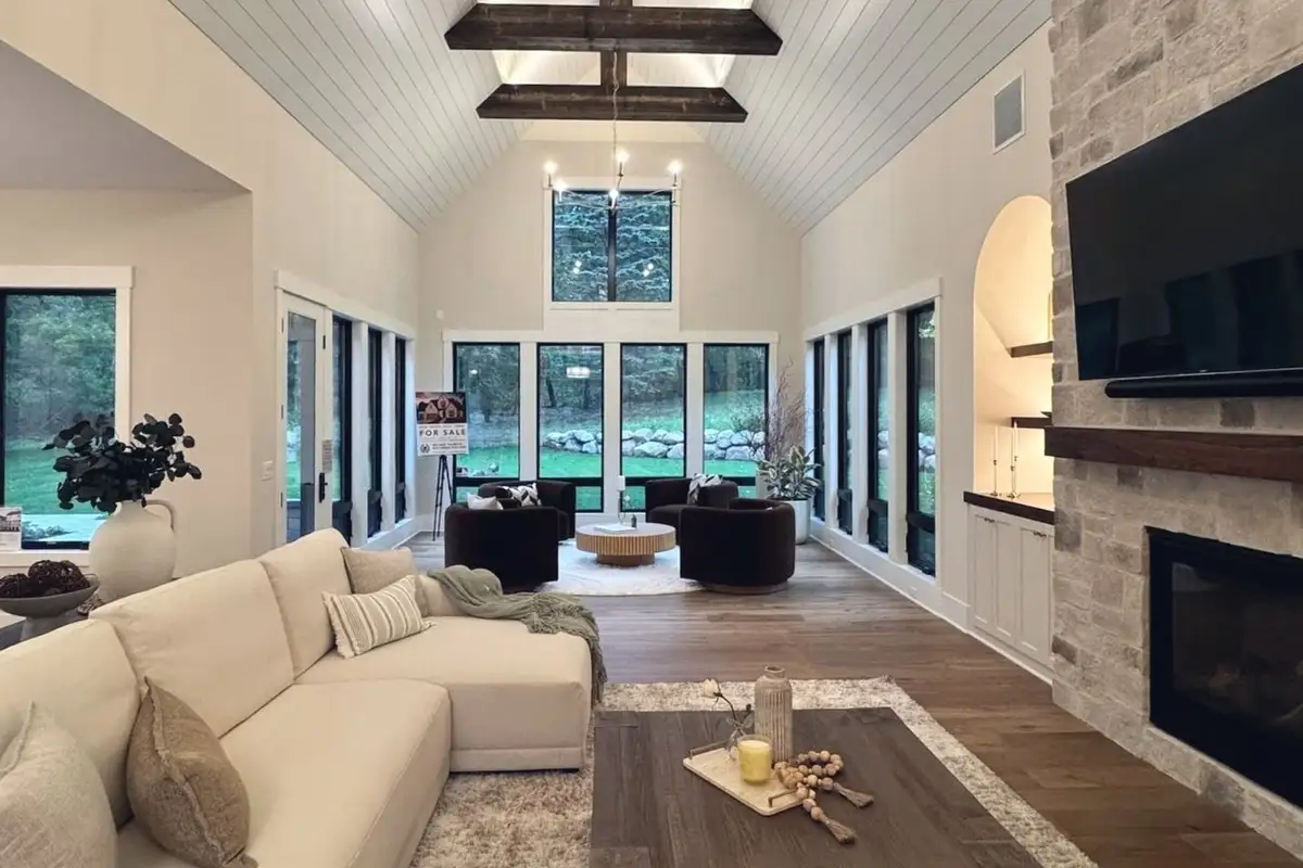 The great room of the TJB Homes model in Edina: a vaulted plank ceiling on dark timber beams, a stacked-stone fireplace and a wall of black-framed windows