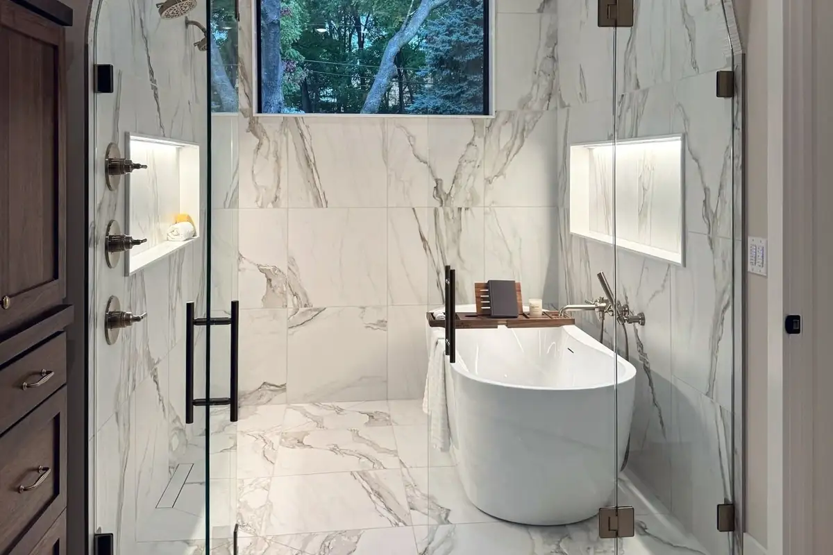 The primary bath of the TJB Homes model in Edina: an arched glass wet room in marble-look tile around a freestanding tub