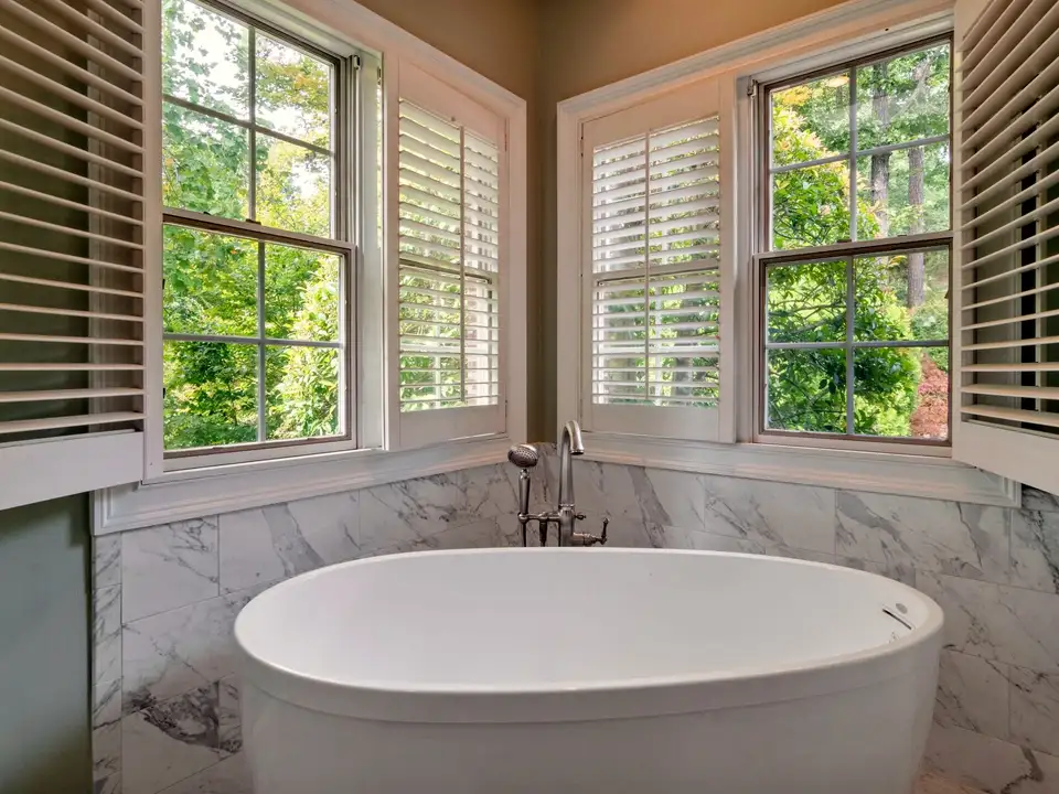 A soaking tub in a shuttered bay window looking into trees