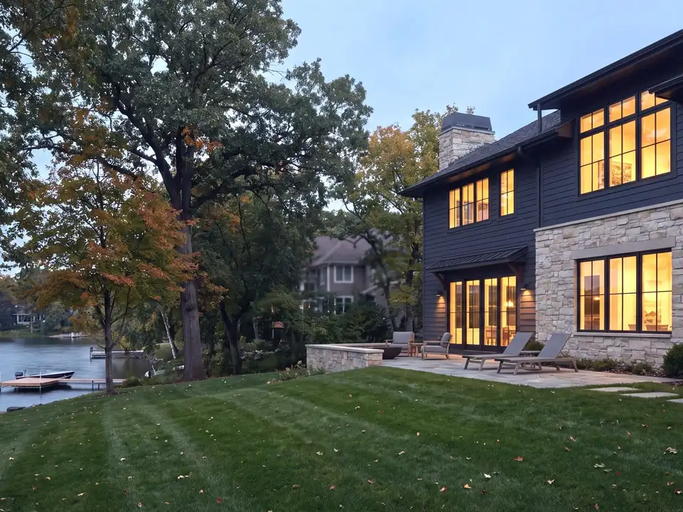 A contemporary lake home of stone and painted siding at dusk, its windows lit, above a lawn and dock
