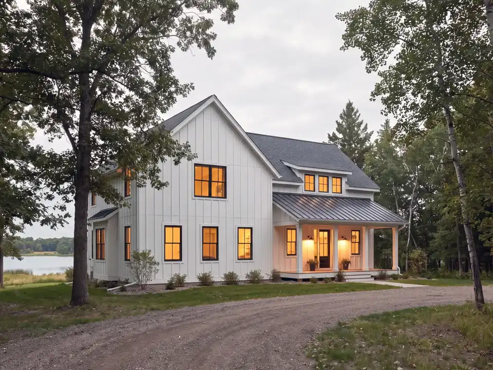 A white board-and-batten modern farmhouse with black windows and a metal porch roof on a wooded lot at evening