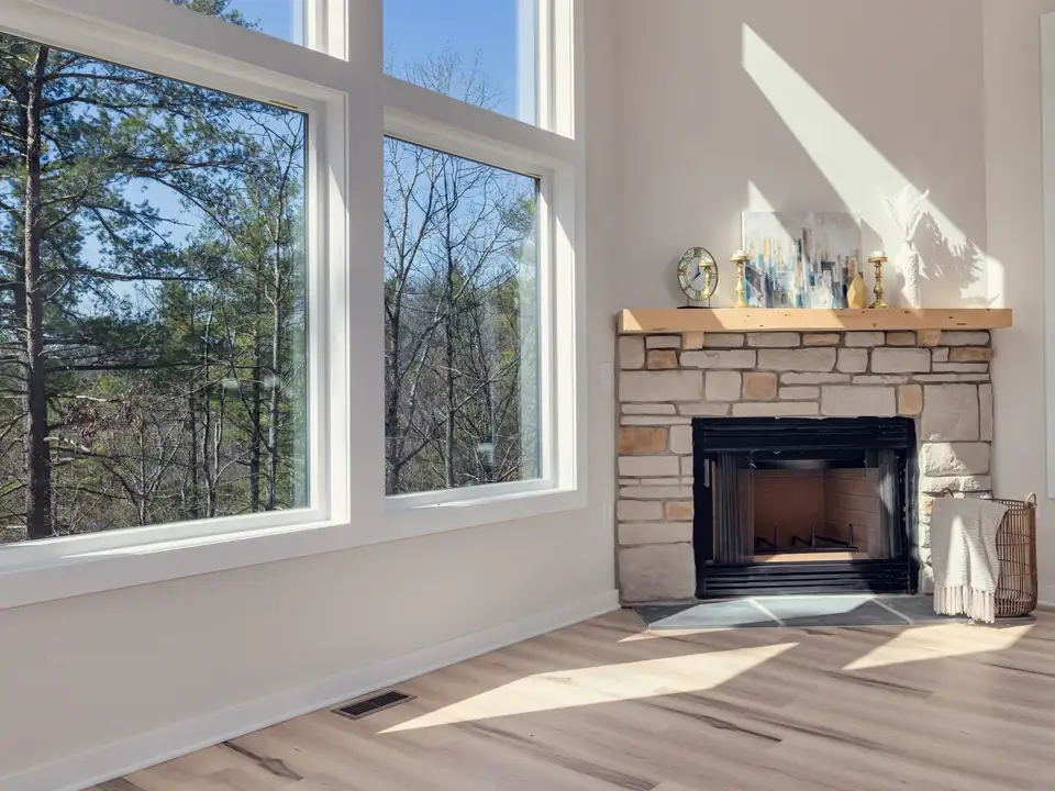 A living room with a stone fireplace and tall windows onto pines