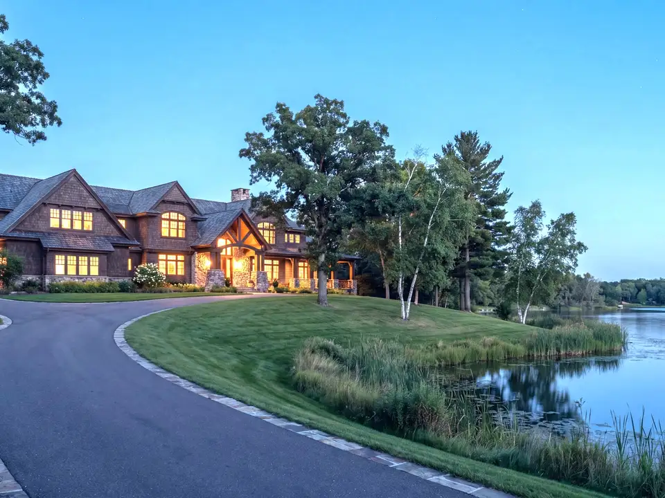 A large shingle-style estate at dusk above a curved drive, its lawn running down to a lake
